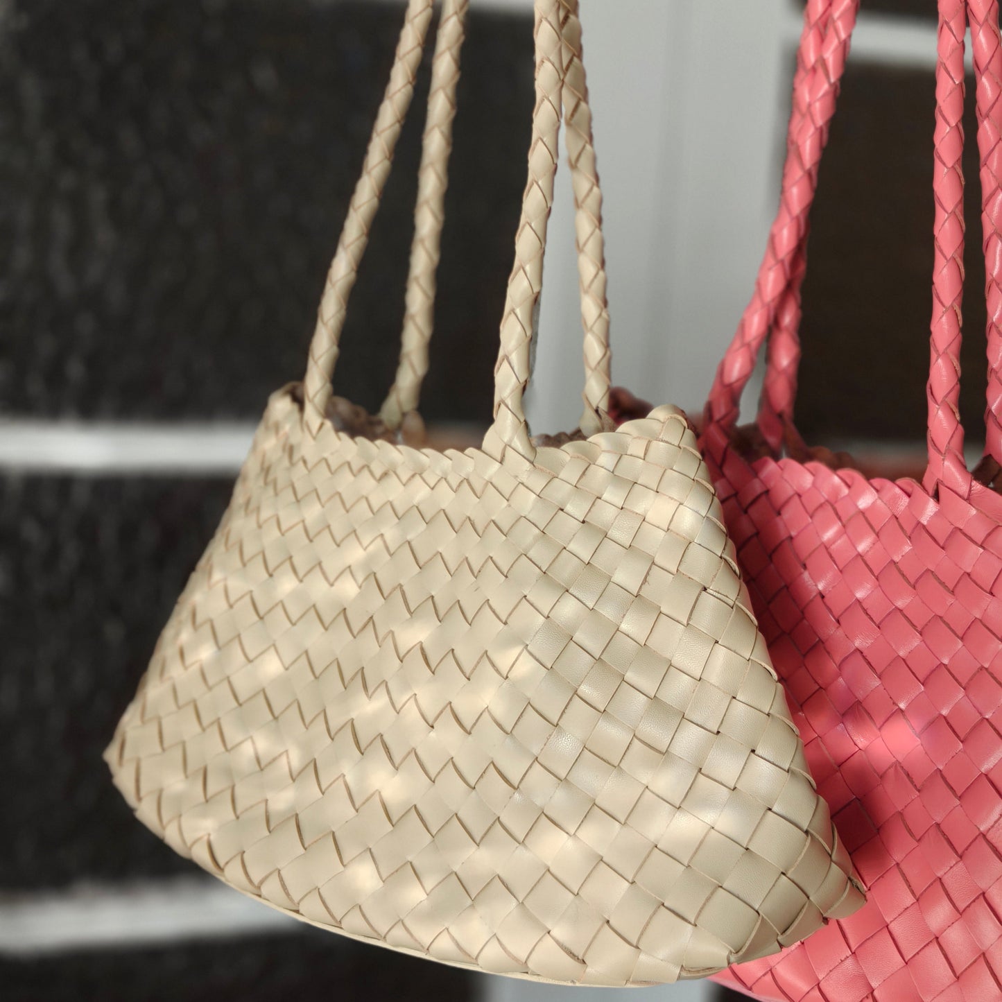 Two woven handbags, one beige and one pink, hanging against a dark background.