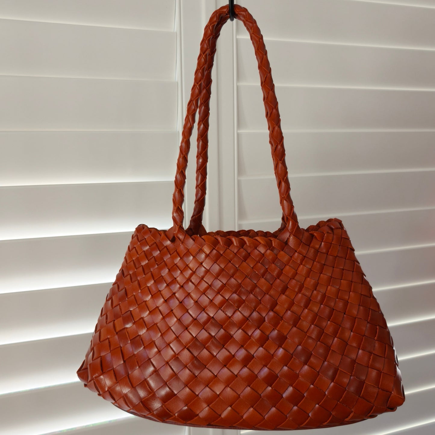 Brown woven handbag hanging against a white window blind background
