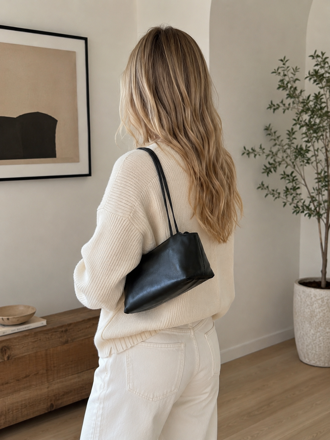 Woman in a beige sweater and white pants holding a black handbag in a room with a plant and wooden bench.