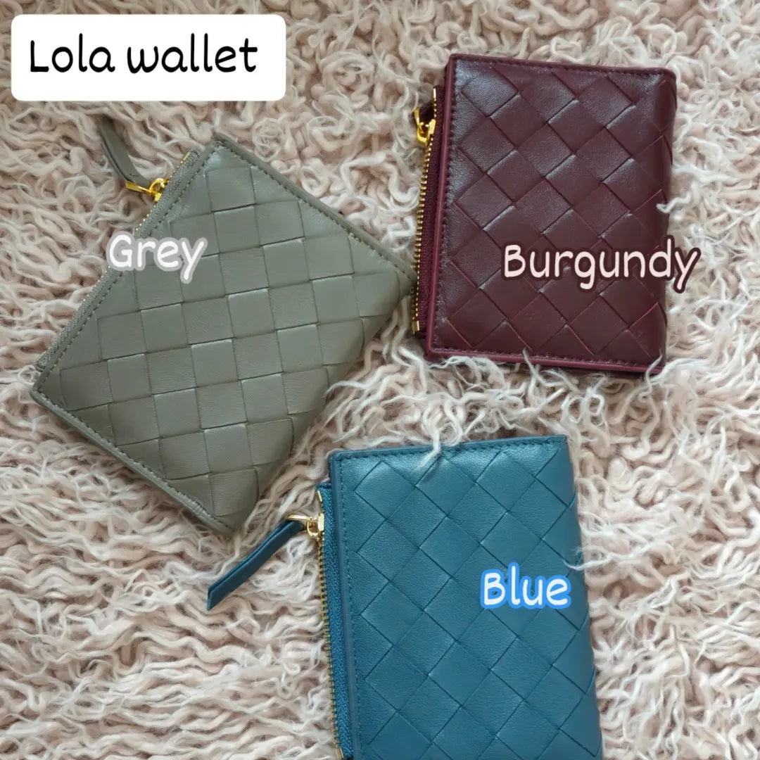 Three Lola sheepskin woven short wallets in grey, burgundy, and blue colors placed on a textured beige surface.