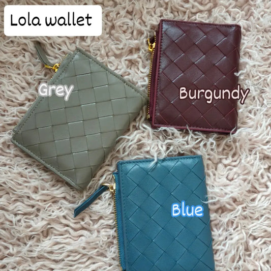 Three Lola sheepskin woven short wallets in grey, burgundy, and blue colors placed on a textured beige surface.